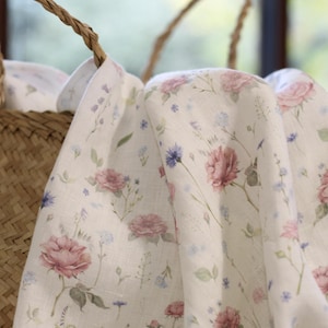 May include: White linen fabric with a delicate floral print featuring pink roses and blue wildflowers. The fabric is draped over a woven basket.