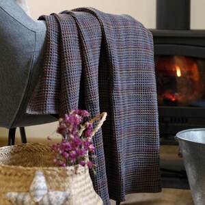 Puede incluir: Una manta tejida en tonos azules, marrones y rojos está sobre un sillón gris. Una cesta de mimbre con flores moradas y una almohada en forma de estrella está frente a la silla. Una chimenea está en el fondo.