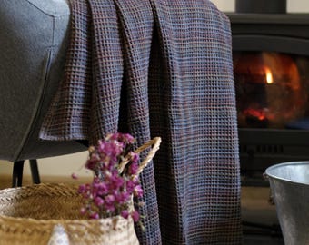 May include: A woven throw blanket in shades of blue, brown, and red is draped over a gray armchair. A wicker basket with purple flowers and a star-shaped pillow sits in front of the chair. A fireplace is in the background.