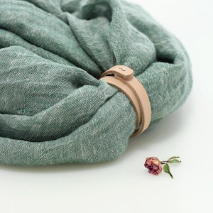 May include: A green linen scarf with a brown leather strap to hold it in place. The scarf is folded and sitting on a white surface.