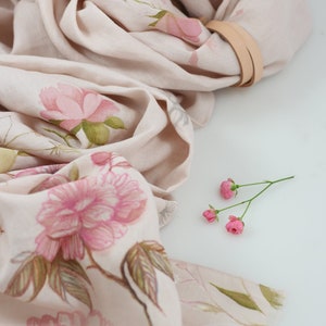 May include: A beige linen scarf with a floral print of pink flowers and green leaves. The scarf is folded and draped over a white surface.