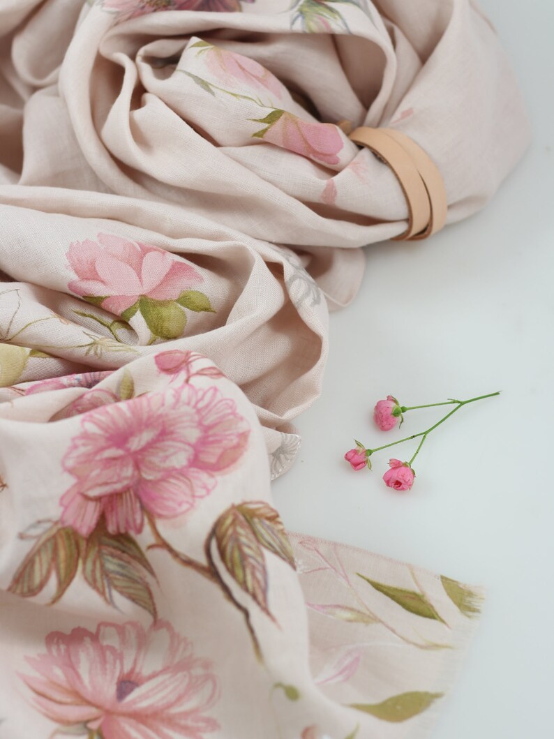 May include: A beige linen scarf with a floral print of pink flowers and green leaves. The scarf is folded and draped over a white surface.