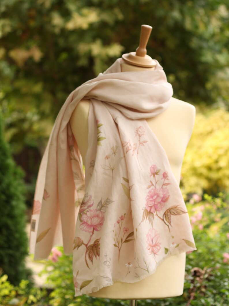 May include: A pale pink scarf with a floral print featuring pink flowers and green leaves. The scarf is draped over a white mannequin.