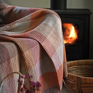 May include: A waffle-weave blanket in shades of orange, pink, and grey, draped over a chair. A fireplace with a burning fire and a woven basket are in the background, creating a warm and inviting atmosphere.