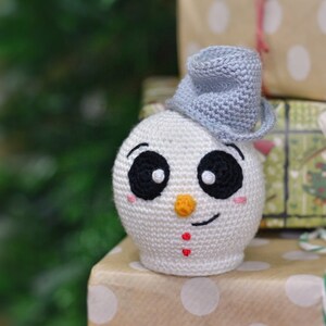 Op de afbeelding: Een handgemaakte, gehaakte sneeuwpop met een blauwe hoed, zwarte ogen en een oranje neus. De sneeuwpop staat op een cadeauverpakking verpakt in bruin papier met witte stippen. De achtergrond is wazig.