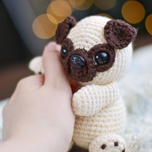 May include: A handmade crochet pug dog with a cream-colored body and dark brown accents around the eyes, ears, and nose. The pug has large black eyes and a smiling face. The toy is held in a person's hand.