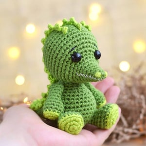 May include: A handmade green crochet alligator toy with black safety eyes and a white stitched mouth. The alligator has a textured body, a row of spikes down its back, and lighter green accents on its feet. The toy is held in a person's hand.