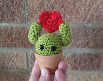 Super cute green crochet  cactus plushies holding a crochet heart above his head