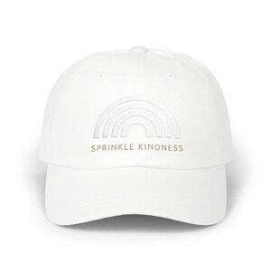 Op de afbeelding: Witte baseballpet met een gebogen klep. De pet heeft een lichtgrijs regenboogontwerp en de tekst "SPRINKLE KINDNESS" in een bruin lettertype. De hoed is gemaakt van een zacht materiaal en is geschikt voor dagelijks gebruik.