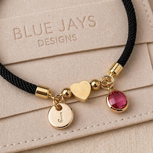 Può includere: Bracciale in corda nera con dettagli dorati, un ciondolo a forma di cuore, un ciondolo rotondo con la lettera "J" e una gemma rosa. Il bracciale è presentato su una carta beige con il testo "BLUE JAYS DESIGNS".