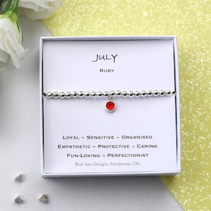 Op de afbeelding: Een zilveren armband met een rode edelsteenbedel. De armband zit in een witte doos met de tekst "JULY RUBY" en een lijst met persoonlijkheidstrekken die geassocieerd worden met de maand juli. De tekst bevat ook "Blue Jays Designs, Handmade Gifts".