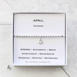May include: A silver bracelet with a charm in the shape of a circle. The bracelet is in a white box with the words "APRIL DIAMOND" and a list of personality traits associated with the month of April: "STRONG - SUCCESSFUL - BRAVE - AFFECTIONATE - ENCOURAGING - GENEROUS - SPIRITED - DETERMINED". The box also includes the text "Blue Jays Designs, Handmade Gifts".