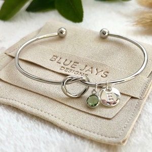 A silver bangle bracelet with a knot in the centre and two charms to the side: a birthstone and an initial disc. The bracelet sits on a branded Blue Jays Designs faux suede pouch.
