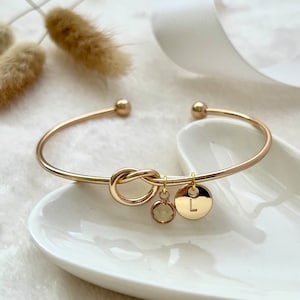 A smooth, yellow-gold bangle with an infinity knot in the centre. A dainty birthstone and matching gold initial disc dangle from one side.  The bangle is set upon a trinket dish and there is dried pampas grass and white ribbon in the background.