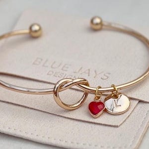 May include: Gold-tone bangle bracelet with a knot design and two charms: a red heart and a circular charm with the letter "L". The bracelet rests on a beige pouch with the text "BLUE JAYS DESIGN".