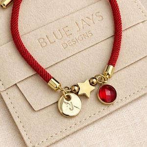May include: A red cord bracelet with a gold-toned adjustable slider, featuring a gold-toned star charm, a circular charm with the letter J, and a round red faceted stone charm. The bracelet is displayed on a beige pouch labeled BLUE JAYS DESIGNS.