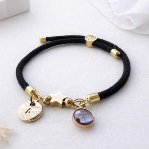 Op de afbeelding: Zwart touw armband met goudkleurige accenten en bedels. De bedels zijn onder meer een ster, een ronde bedel met de letter "A" en een paarse edelsteen. Een witte geschenkdoos staat op de achtergrond.