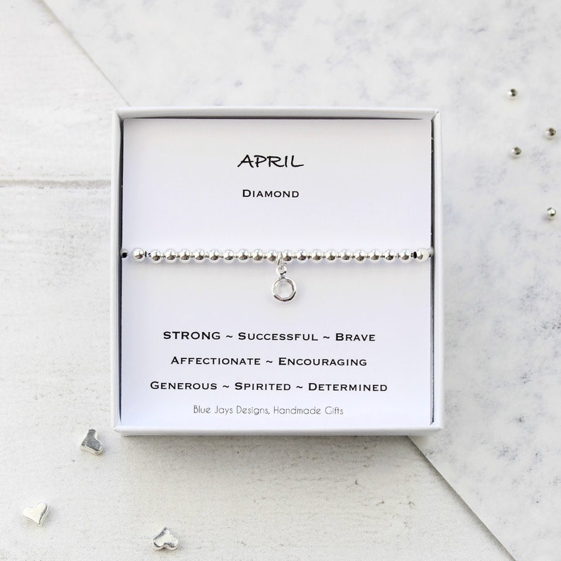 May include: A silver bracelet with a charm in the shape of a circle. The bracelet is in a white box with the words "APRIL DIAMOND" and a list of personality traits associated with the month of April: "STRONG - SUCCESSFUL - BRAVE - AFFECTIONATE - ENCOURAGING - GENEROUS - SPIRITED - DETERMINED". The box also includes the text "Blue Jays Designs, Handmade Gifts".