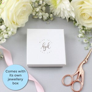 May include: A white box with a logo that says "Blue Jays Designs byd" in brown lettering. The box is sitting on a gray surface with white flowers and a pink ribbon.