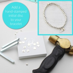 May include: A silver bracelet with a small silver disc charm with the letter "E" stamped on it. The bracelet is on a white surface with a hammer and a box of silver discs with letters stamped on them. The text "Add a hand-stamped initial disc to your bracelet" is on a blue circle.
