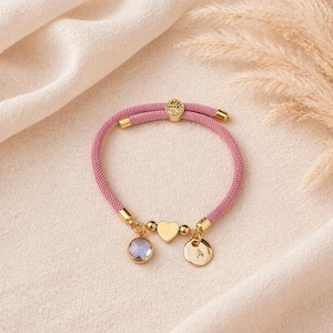 Soft pink cord bracelet with heart charm, engraved initial disc and birthstone on neutral background