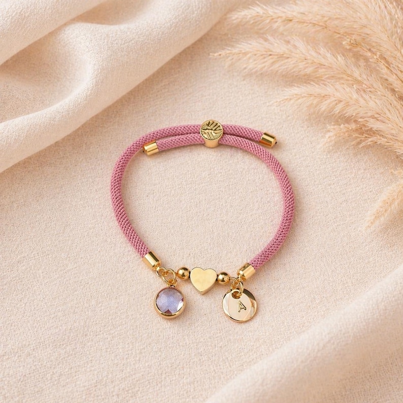 Soft pink cord bracelet with heart charm, engraved initial disc and birthstone on neutral background