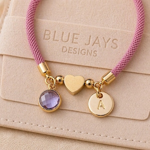Può includere: Un braccialetto rosa intrecciato con dettagli dorati e ciondoli. I ciondoli includono un cuore, un ciondolo rotondo con la lettera "A" e una gemma viola sfaccettata. Il testo "BLUE JAYS DESIGNS" è visibile sullo sfondo.