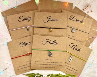 May include: A set of eight personalized friendship bracelets with charms, each packaged in a brown paper envelope with the recipient's name written in cursive. The charms include a bell, a snowflake, a star, a gingerbread man, a reindeer, and a Christmas tree. The bracelets are tied with colorful string and have the words "Make a Wish" printed on the packaging.