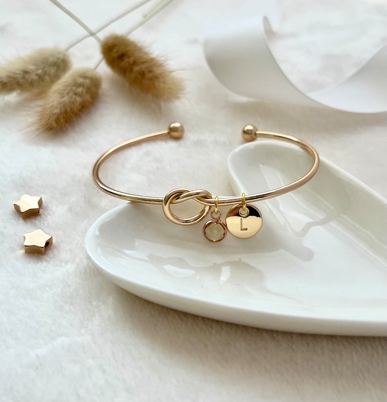 A smooth, yellow-gold bangle with an infinity knot in the centre. A dainty birthstone and matching gold initial disc dangle from one side.  The bangle is set upon a trinket dish and there is dried pampas grass and white ribbon in the background.