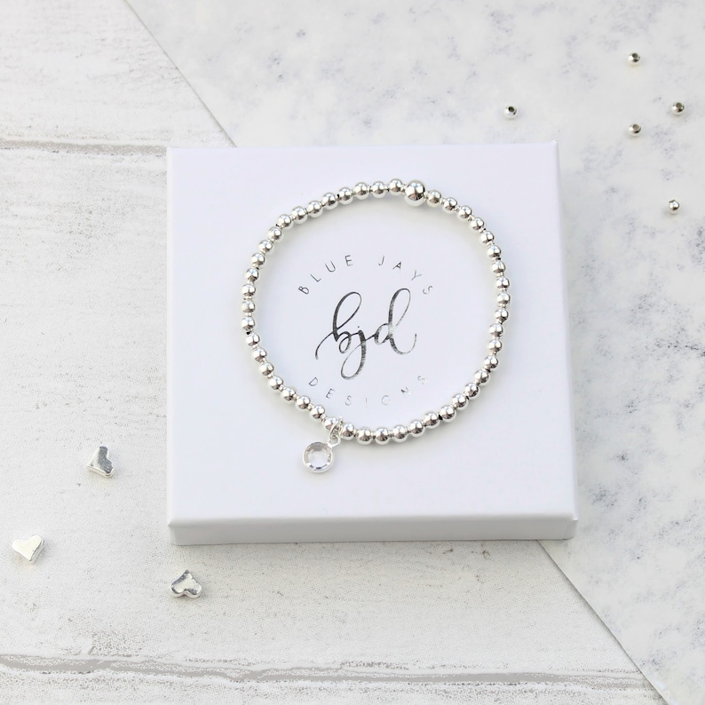 May include: A silver bracelet with a clear crystal charm. The bracelet is in a white box with the text "Blue Jays byd Designs" on it.