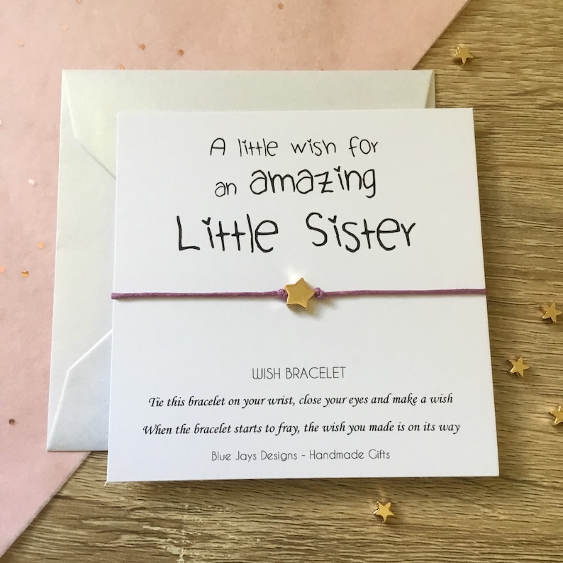May include: A white card with the text "A little wish for an amazing Little Sister" and a purple string bracelet with a gold star charm. The card also includes the text "WISH BRACELET" and instructions for making a wish.