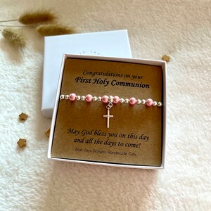 May include: A white gift box with a brown card inside. The card has the text "Congratulations on your First Holy Communion" and "May God bless you on this day and all the days to come!"  The card also has the text "Blue Jays Designs, Handmade Gifts". A pink and silver beaded bracelet with a silver cross charm is on top of the card.