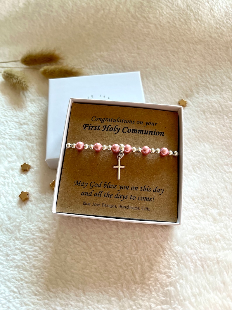 May include: A white gift box with a brown card inside. The card has the text "Congratulations on your First Holy Communion" and "May God bless you on this day and all the days to come!"  The card also has the text "Blue Jays Designs, Handmade Gifts". A pink and silver beaded bracelet with a silver cross charm is on top of the card.