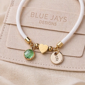 Pode incluir: Pulseira de cordão branco com berloques dourados, incluindo uma gema verde, um coração e um disco com a letra "S". A pulseira é apresentada em um fundo bege com o texto "BLUE JAYS DESIGNS".