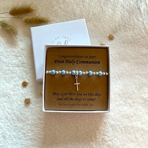 May include: A white gift box with a brown insert that says "Congratulations on your First Holy Communion". Inside the box is a blue and silver beaded bracelet with a silver cross charm. The text on the insert also says "May God bless you on this day and all the days to come!" and "Blue Jays Designs, Handmade Gifts".