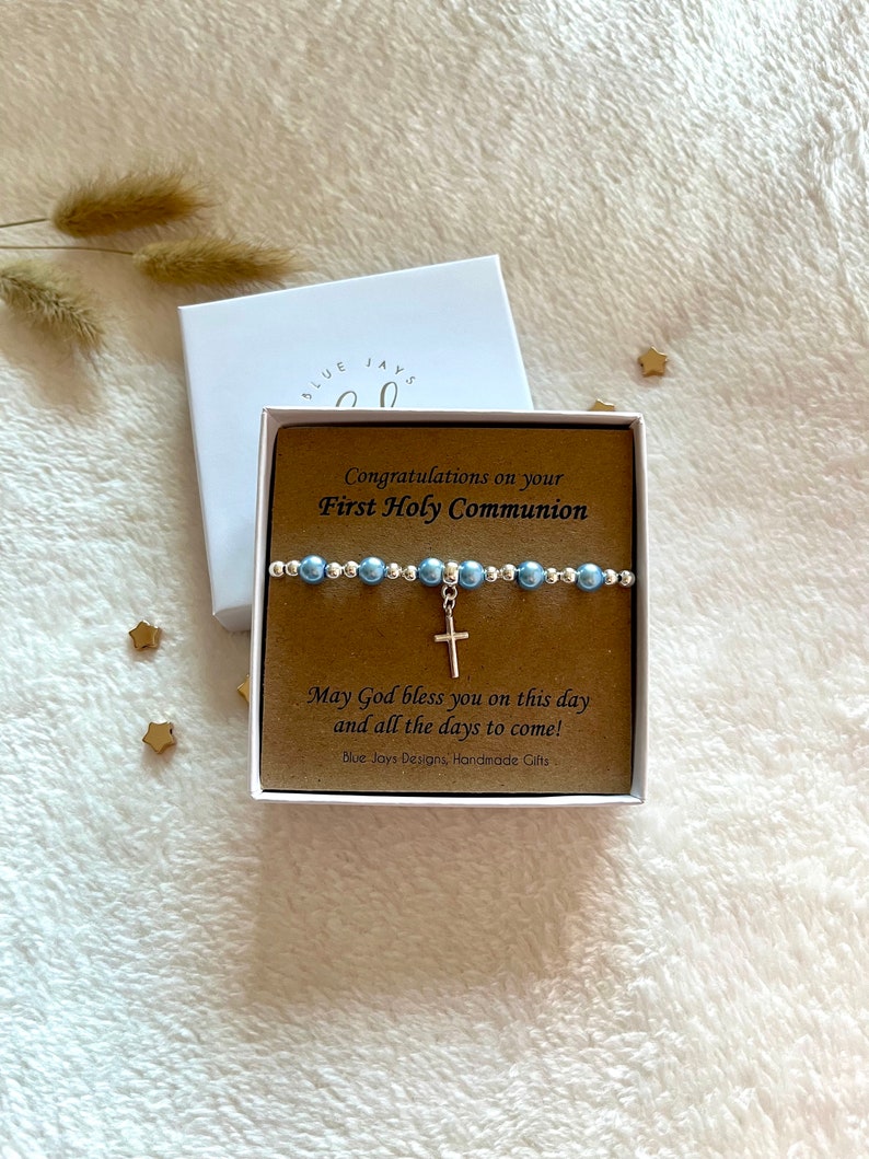 May include: A white gift box with a brown insert that says "Congratulations on your First Holy Communion". Inside the box is a blue and silver beaded bracelet with a silver cross charm. The text on the insert also says "May God bless you on this day and all the days to come!" and "Blue Jays Designs, Handmade Gifts".