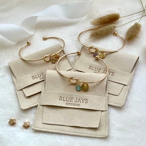 May include: Gold-toned bracelets with knot details and charm pendants, displayed on beige pouches. The charms include gemstones and initial letters. The pouches are embossed with "BLUE JAYS DESIGNS". Two gold star-shaped earrings are also visible.