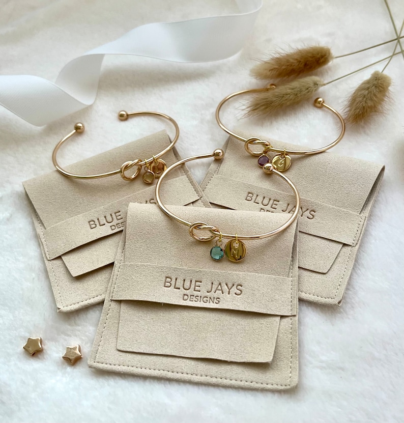 May include: Gold-toned bracelets with knot details and charm pendants, displayed on beige pouches. The charms include gemstones and initial letters. The pouches are embossed with "BLUE JAYS DESIGNS". Two gold star-shaped earrings are also visible.