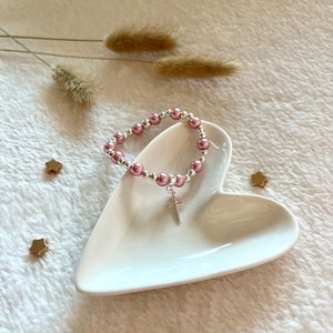 May include: A pink and silver beaded bracelet with a silver cross charm. The bracelet is displayed on a white heart-shaped dish.