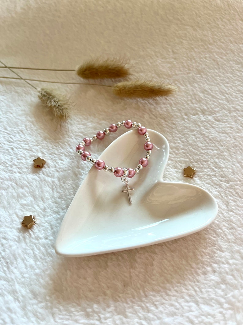 May include: A pink and silver beaded bracelet with a silver cross charm. The bracelet is displayed on a white heart-shaped dish.