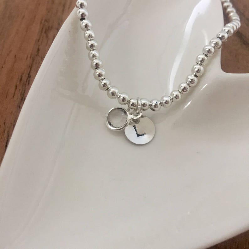 May include: A silver beaded bracelet with a small silver charm featuring the letter "L" and a clear stone.