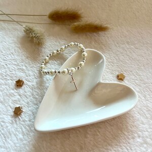 May include: A white pearl bracelet with a silver cross charm on a white heart-shaped dish. The bracelet is on a white fuzzy surface with gold star-shaped accents.