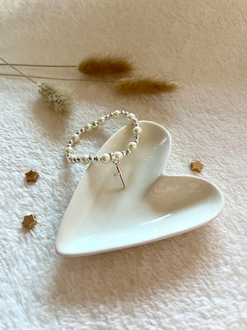 May include: A white pearl bracelet with a silver cross charm on a white heart-shaped dish. The bracelet is on a white fuzzy surface with gold star-shaped accents.