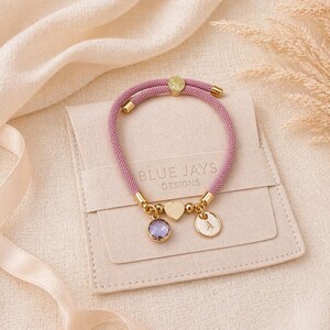Pink cord personalised heart bracelet with initial charm and birthstone displayed on cream gift pouch