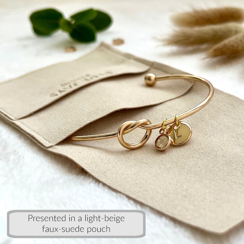 May include: Gold-colored bangle bracelet with a knot design and two charms, one with the letter "L". The bracelet is presented in a light-beige faux-suede pouch. The pouch has the text "SAVE JAYS" printed on it.