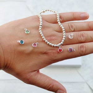 May include: A silver bead bracelet with a variety of colored gemstone charms. The charms are in shades of red, pink, green, blue, and purple. The bracelet is on a hand, and the charms are scattered around it.