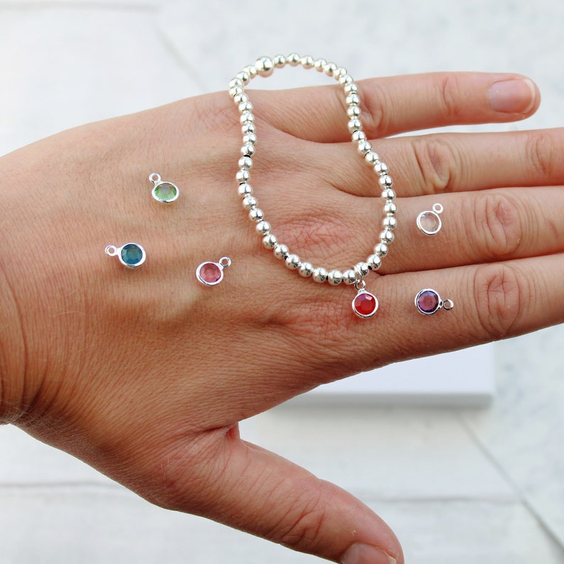 May include: A silver bead bracelet with a variety of colored gemstone charms. The charms are in shades of red, pink, green, blue, and purple. The bracelet is on a hand, and the charms are scattered around it.