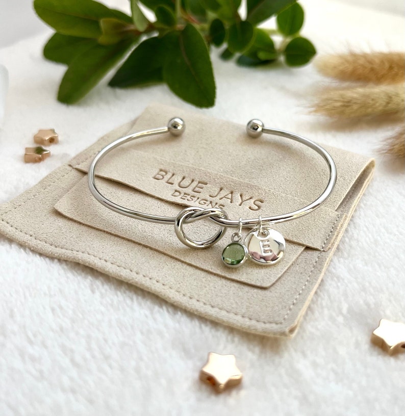 A silver bangle bracelet with a knot in the centre and two charms to the side: a birthstone and an initial disc. The bracelet sits on a branded Blue Jays Designs faux suede pouch.