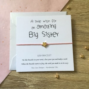 May include: A white card with pink string tied around it. The card says "A little wish for an amazing Big Sister". The string has a gold star charm. The card also says "WISH BRACELET" and "Tie this bracelet on your wrist, close your eyes and make a wish. When the bracelet starts to fray, the wish you made is on its way. Blue Jays Designs - Handmade Gifts."