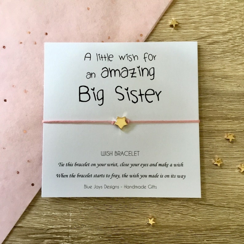 May include: A pink and white card with the text "A little wish for an amazing Big Sister" and a pink string bracelet with a gold star charm. The card also includes instructions for making a wish on the bracelet.